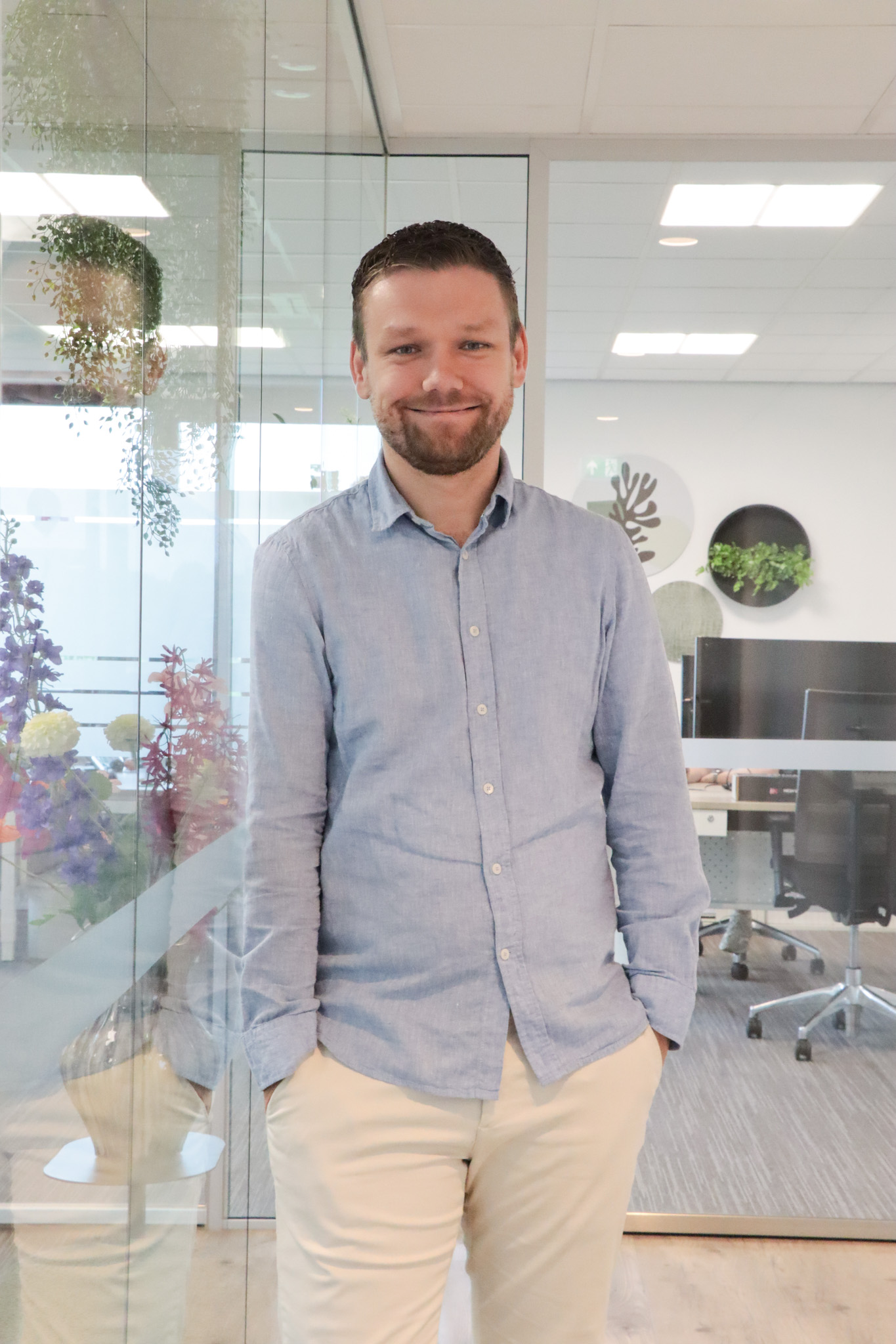 Smiling man in a light blue button-down shirt and beige pants standing in a glass-walled office space with plants in the background.