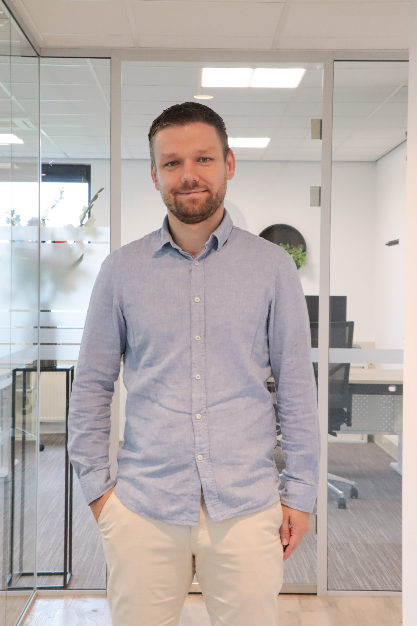 Man standing in a bright office wearing a light blue button-down shirt and beige pants, with glass partitions behind him.