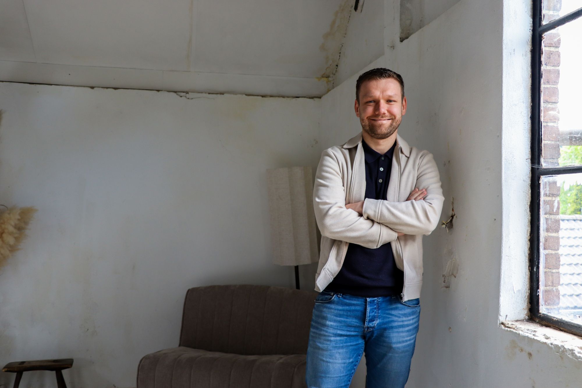 Smiling man with arms crossed stands in a bare, unfinished room next to a large window on the right.