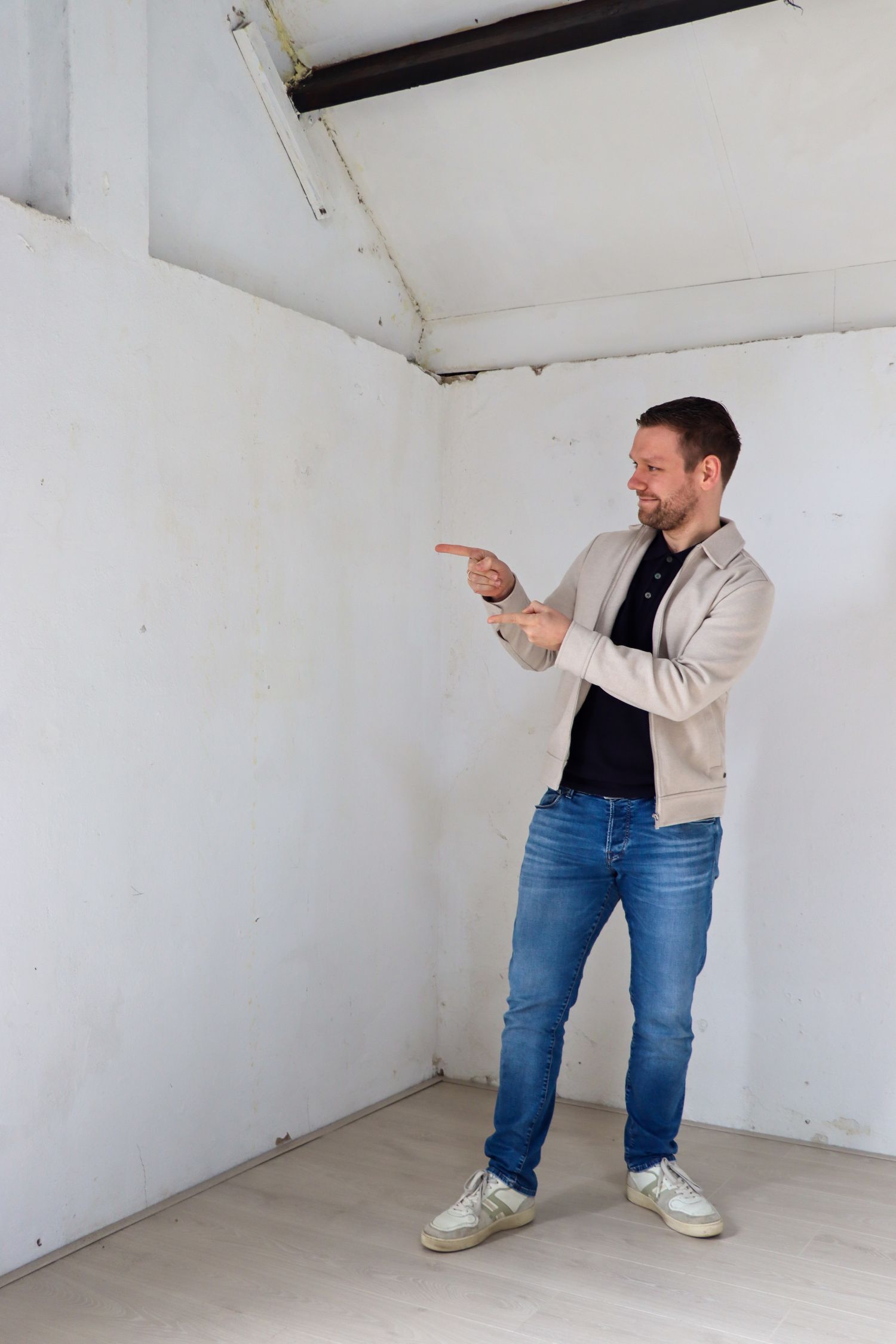 Man in a beige jacket and jeans stands in an empty white-walled room, pointing to the left with both hands as if indicating something off screen.